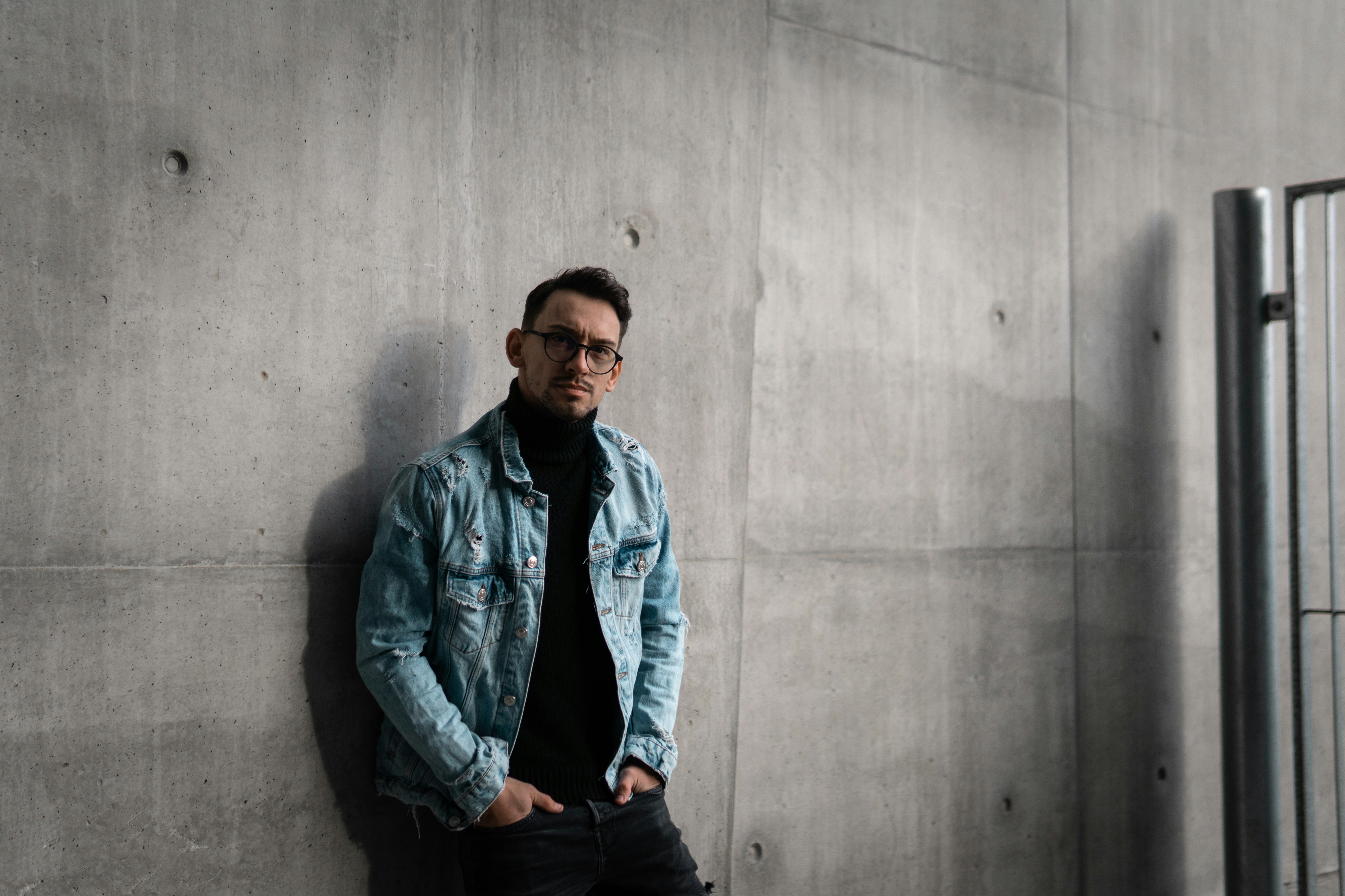 man in blue denim jacket and black pants standing beside gray wall